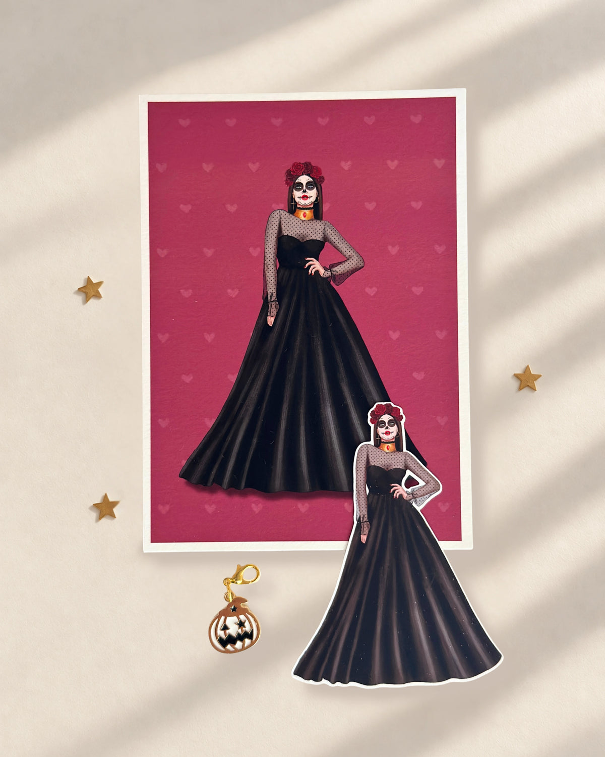 Pack de Halloween La Catrina con postal ilustrada de una catrina con vestido negro y corona de rosas rojas, sticker troquelado a juego y charm metálico de calabaza en blanco, negro y dorado.