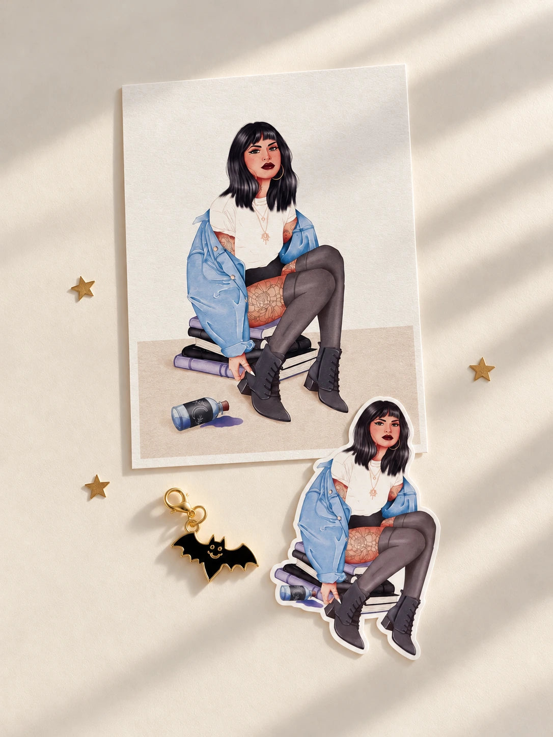 Pack de papelería ilustrada de Halloween con postal y sticker de una chica de pelo negro, chaqueta vaquera azul, medias oscuras y botas negras, sentada sobre una pila de libros, junto a un charm metálico de murciélago negro con detalles dorados. Presentación sobre fondo crema y estrellas doradas.