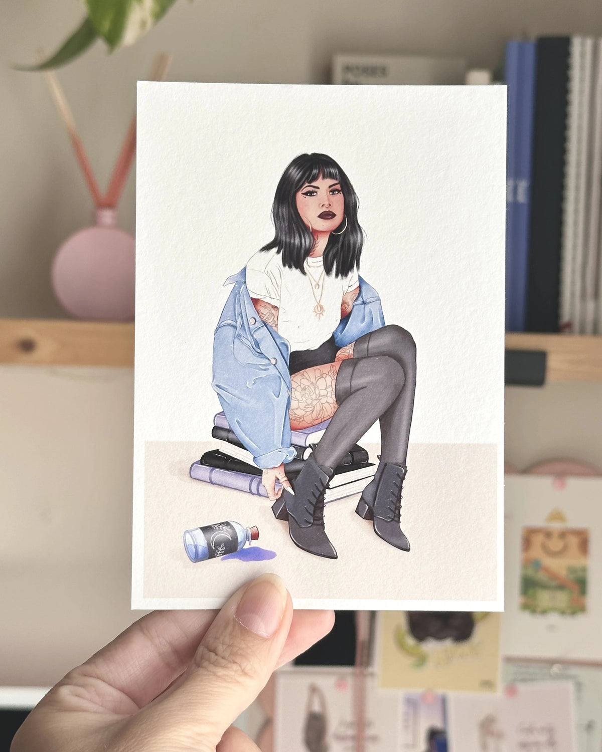 Postal ilustrada Brujita Moderna de Natalia Naya, con una chica de pelo negro, chaqueta vaquera azul, tatuajes y botas negras, sostenida con la mano frente a un escritorio creativo.