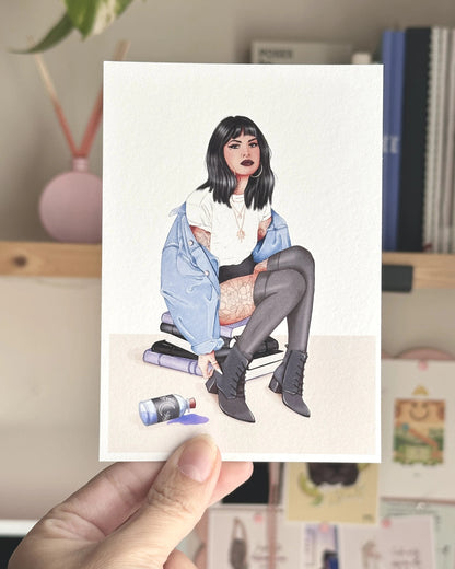 Postal ilustrada Brujita Moderna de Natalia Naya, con una chica de pelo negro, chaqueta vaquera azul, tatuajes y botas negras, sostenida con la mano frente a un escritorio creativo.
