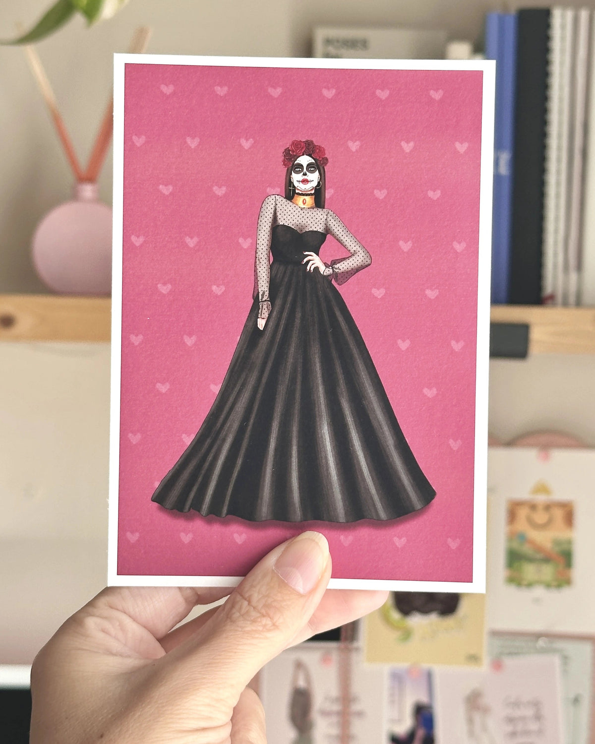 Postal ilustrada de La Catrina de Natalia Naya, con una mujer de vestido negro largo, corona de rosas rojas y maquillaje de calavera mexicana sobre fondo fucsia.
