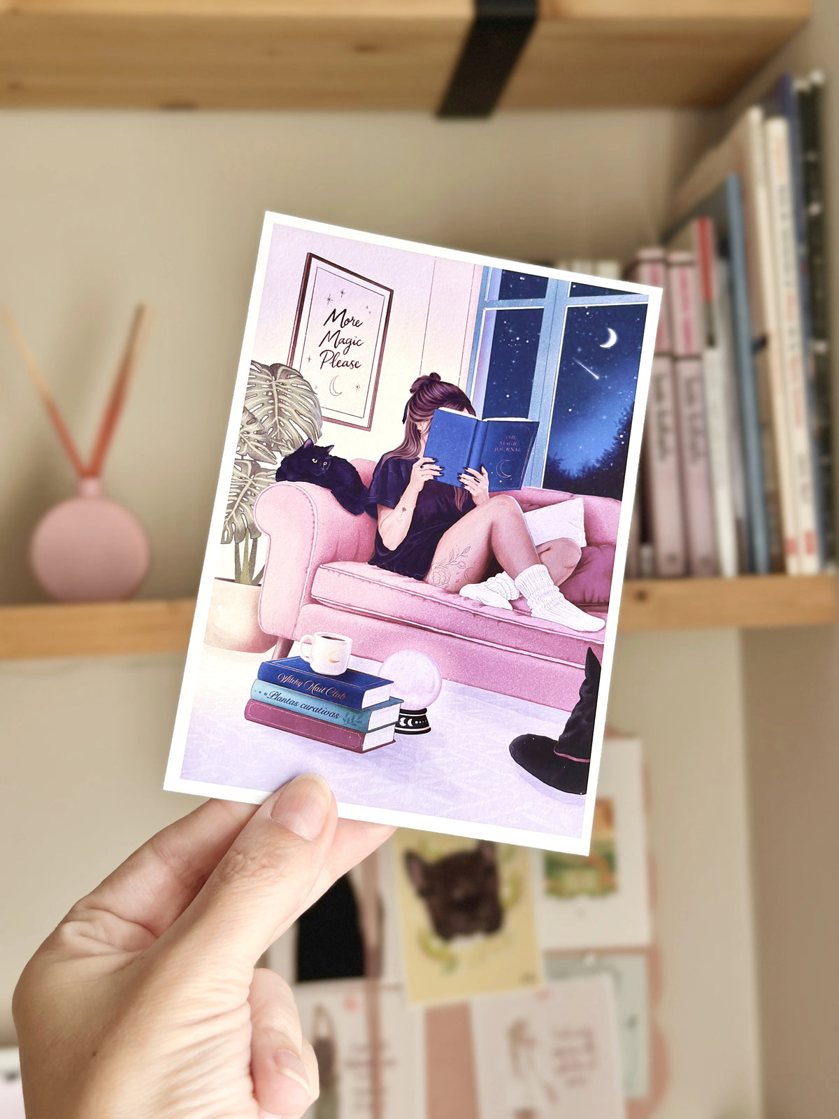 Detalle de una postal ilustrada de una chica leyendo en un sofá rosa, impresa en papel con borde blanco.
