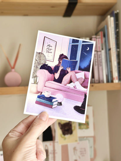Detalle de una postal ilustrada de una chica leyendo en un sofá rosa, impresa en papel con borde blanco.